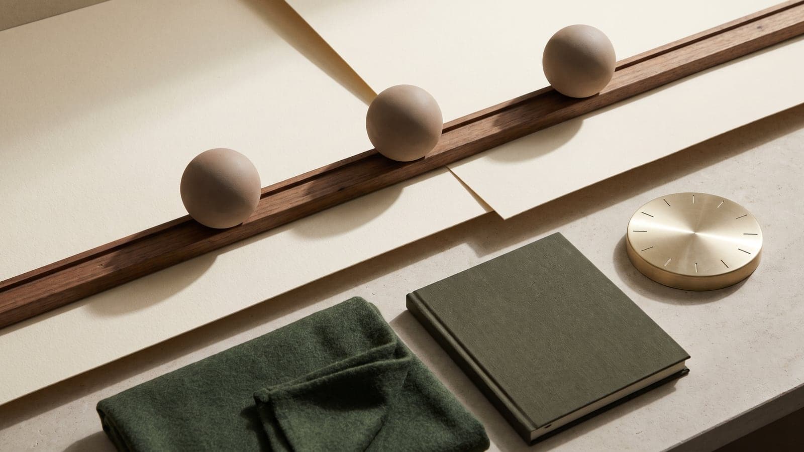 Three brown spheres rest in a grooved wood rail on layered ivory paper, next to a green book, a green cloth, and a brass dial.