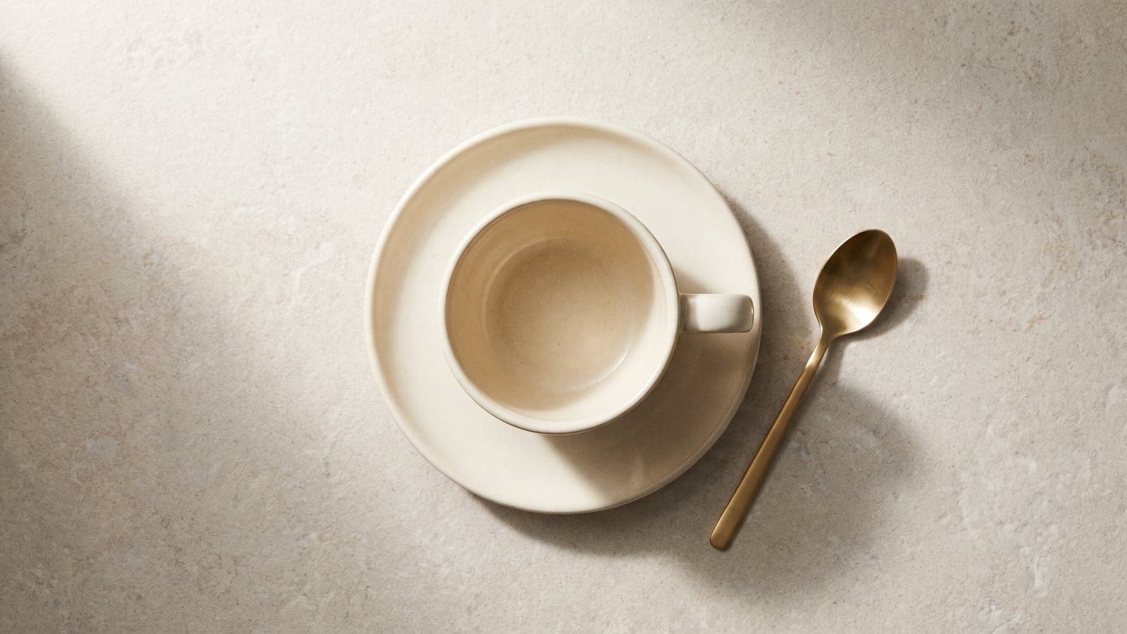 A single, well-crafted ceramic cup on an empty saucer viewed from above, on pale limestone, one soft shadow.