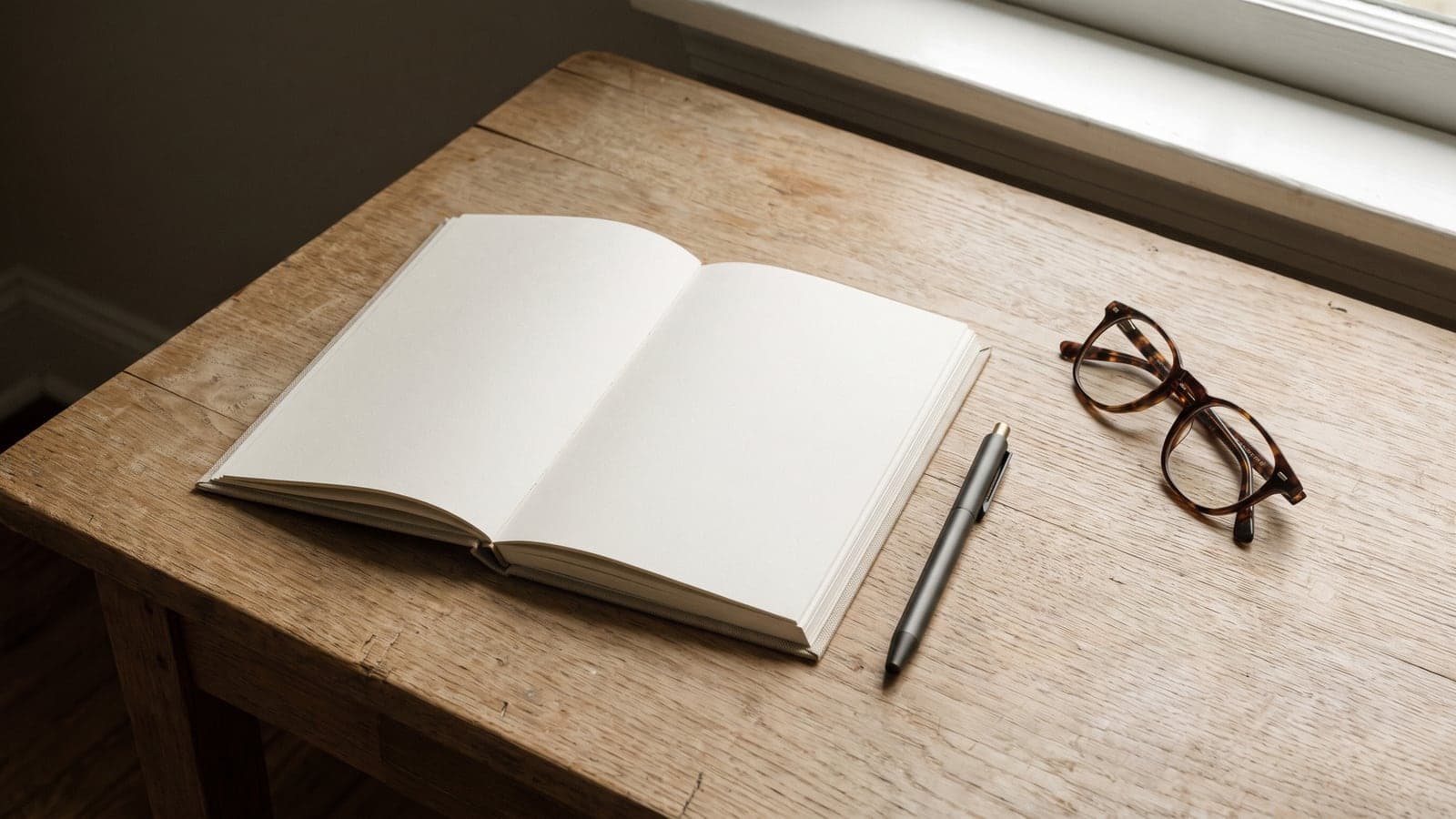 A close-up of a clean, empty notepad and a simple pen on a light oak surface next to a pair of glasses, soft window light.