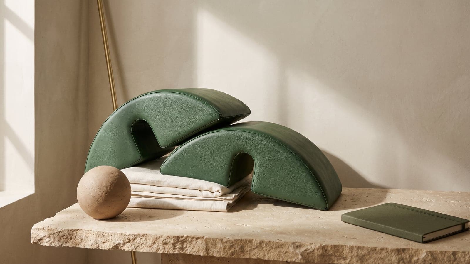 Two green arc-shaped cushions, a clay sphere, and folded ivory linen rest on a rough limestone slab in warm, raking side light.