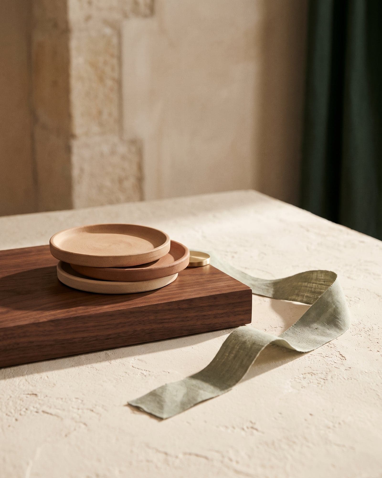 Three offset clay discs stacked on a walnut plinth with a sage ribbon across ivory plaster